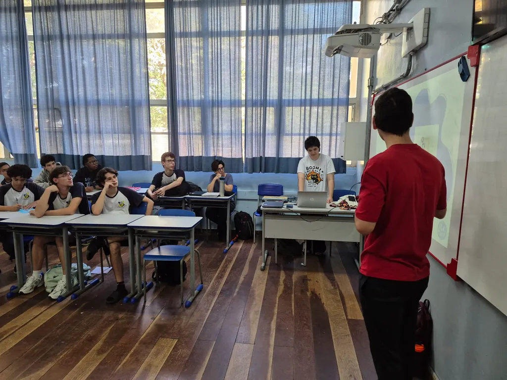 Fotografia retangular colorida. Em primeiro plano, um membro do Olá, Mundos! leciona uma parte da apresentação para os alunos da Escola Estadual Vitor Meirelles, em uma sala de aula. Ao fundo, à esquerda, observam-se oito estudantes prestando atenção à esquerda. À direita, outro membro da entidade acompanha a apresentação.