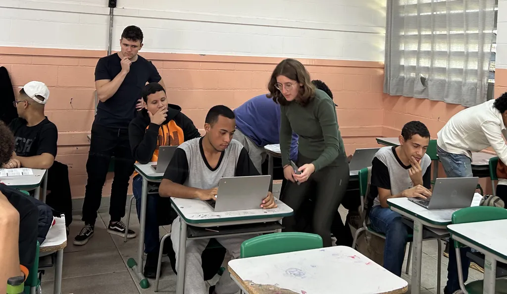Imagem de uma aula do Olá, Mundos! Em várias carteiras, alunos concentrandos mexendo em laptops, sendo auxiliados por membros do projeto.